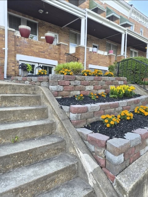 Brick building facade with tiered garden beds containing yellow flowers and dark mulch, concrete steps in foreground