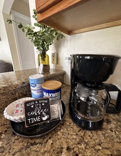Kitchen counter with coffee maker, Maxwell House coffee, granulated sugar, coffee-themed sign, plants in vase, and wooden ceiling beam