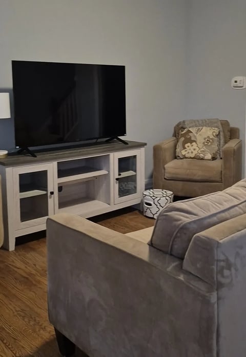 A living room with a gray couch, tan armchair, and white TV stand holding a black flatscreen TV on a wooden floor