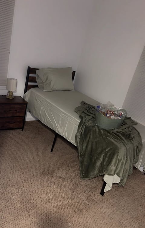 Sparse bedroom with a single bed with cream sheets and pillow, dark wooden nightstand with lamp, green blanket on bed, and beige carpet flooring